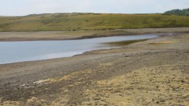  Llyn Aled Isaf in Conwy. The banks of the lake are dried up, the nearest bank is sandy with pebbles on, the bank in the distance is green, with a hill rising up behind it. 