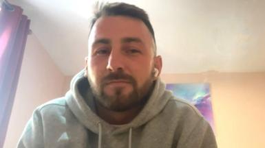 A man with short brown hair and a beard is wearing grey hoodie as he takes a video call from a hotel room