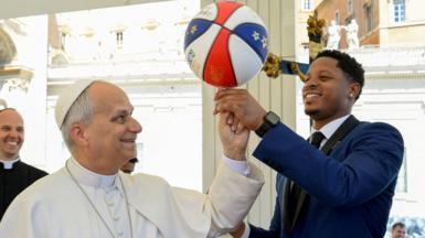 Pope spinning a basketball on his finger with the help of a Harlem Globetrotter player