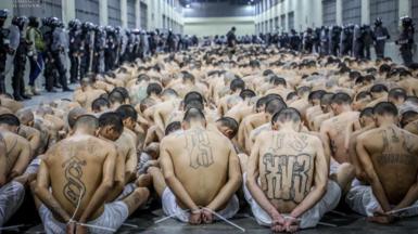 Inmates with their hands tied behind their back and stripped down to their waists arrive at the Cecot prison in Tecoluca on March 15, 2023. They are seated close to each other in long rows in a corridor, while being watched over by guards in riot gear. 