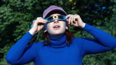 A woman wearing blue looking through special cardboard glasses for looking at the sun