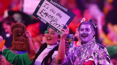 Fans in Wizard of Oz fancy dress - a woman dressed as Dorothy and a man dressed as the Tin Man - cheer at a darts match. The woman is holding a plushy dog toy and a sign saying "is this the Emerald City?" The man is holding a drink.