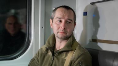 Denys, sitting on a train, looks down away from the camera, lips downturned. He is wearing military fatigues. 