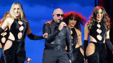 Pitbull, a bald man wearing sunglasses, holds a microphone as he performs on stage next to a group of female dancers.