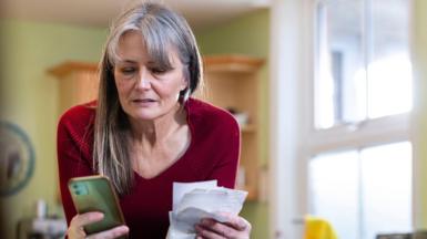 Middle-aged woman with long grey hair looks at a pile of receipts and her mobile phone. She has a slightly anxious look on her face.