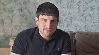 A young man looks at the camera. He has relatively short, dark hair and is wearing a dark polo shirt.