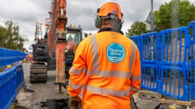 Thames Water worker in orange overalls with Thames Water logo on the back in front of a digger on the road