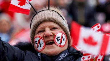A person in a beige woollen hat and navy jacket and with stickers on the face holds a Canadian flag