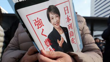 A close up of a pamphlet with a picture of Sanae Takaichi on the front held by a person off camera. Sanae Takaichi is wearing a black suit jacket, white t-shirt and pearl necklace in the photograph on the pamphlet. She is smiling. It has a white background and red border with red writing.