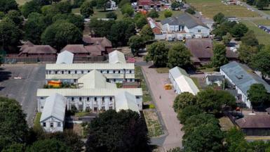 A former military base at Manston, used as a detention and processing centre for migrants who arrived in the UK by small boat - it is seen from overhead, as a series of interlinked white buildings surrounded by fields and trees 