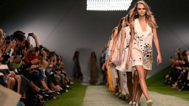 Model Cara Delevingne walks in a straight line of several women along a catwalk, surrounded by crowds either side.