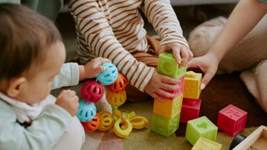 High angle shot of children's hands holding multi-coloured bricks and rattle while adult hand assisting them to build tower