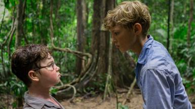 A young boy with brown hair and glasses looks up at a blonde haired boy in a blue shirt. They are in a wooded area of a tropical island.