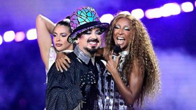 British musician Boy George and Italian singer Senhit Zadik Zadik aka Senhit perform in silver and black sparkly outfits, with blue and purple lights visible behind them.