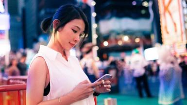 A woman in a white sleeveless top stands in New York City's Times Square at dusk, looking down at the smartphone in her hands