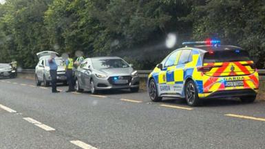 Garda officers and garda cars surround a small car on the roadside of the N2  near Carrickmacross. The officers are all wearing blue uniforms, but two are wearing yellow high-viz vests over their shirts.  The boot of a hatchback vehicle behind them is open. 