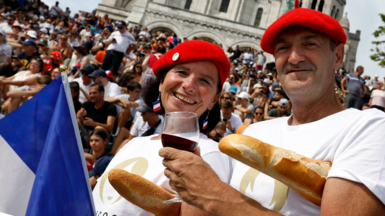 A man and woman wearing French berets hold red wine and baguettes in front of a crowd