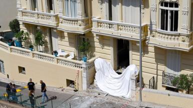 Frontage of a large cream coloured residential building in Monaco with damage to windows and a white sheet accross the front steps and police standing near the front of the building