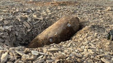 The picture shows a large, rusted cylindrical object, a a German SC250 air-dropped bomb, lying on its side in a shallow pit at what appears to be a construction or excavation site. The object is partially embedded in loose grey rubble made up of broken stone and aggregate.