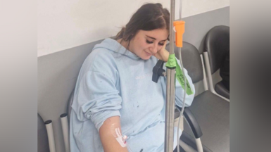 Paige Roberts sitting down in a hospital chair having IV fluids administered. She is wearing a light blue hospital gown and is looking down at the floor