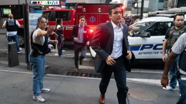 A man runs towards the camera, directed by a female police officer in plain clothes and wearing a tactical vest, with several police and fire vehicles in the background
