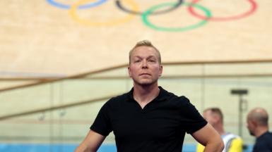 Sir Chris Hoyle, looking calm in a black top, stands below the Olympic rings