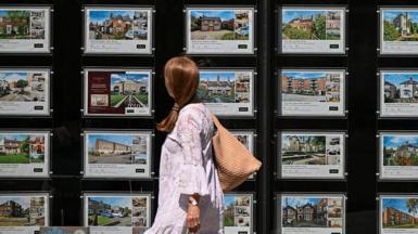 A woman in a white flowy dress walks past an estate agent window filled with home listings.