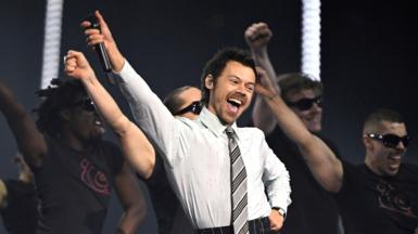 Harry Styles performs at the Brit Awards. He is wearing a grey office shirt and tie combination.