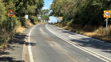 An A road, with road markings. Trees line the road on either side side with 50mph speed limit signs either side.