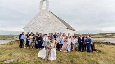 This image shows a couple after getting married, they are stood outside the little church with their wedding guests stood behind them. 