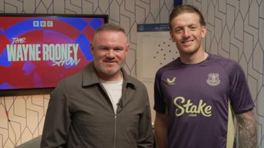 Wayne Rooney and Jordan Pickford pose in front of a TV screen that has an advert for The Wayne Rooney Show on it