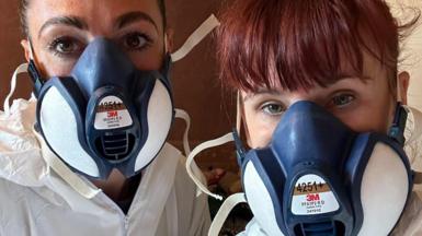 Two women in a selfie. The woman on the right has ginger hair tied back and a fringe. Her colleague has brown hair which has been tied back. They are wearing white hazmats suit and navy face masks.