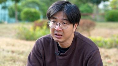 Close up of William Li. He has straight black hair, circular glasses and is wearing a sweater. As he speaks he looks upset.