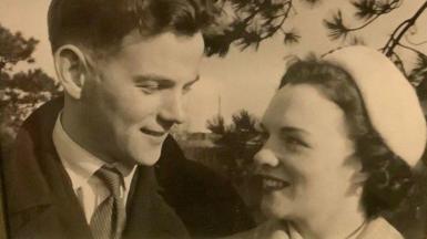 A black and white photo of a young man and woman looking at each other affectionately. The man is wearing an overcoat, a light shirt and a striped tie. The woman has short, curly dark hair under a light hat and is wearing lipstick.