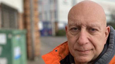 Dave wears a bright orange and grey work anorak and a grey hoodie. He is bald and stands next to the road outside the Vauxhall factory. The Vauxhall emblem is on one corner, and the building recedes into the distance with many windows across its two storeys.. 