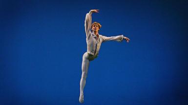 Russian ballet star Vladimir Shklyarov poses in a ballet pose, wearing a white and gold costume. His head is tilted back and his eyes are closed. The background is dark blue.