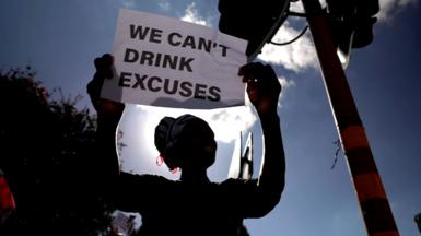 A woman is seen silhouetted against a blue sky with a sign reading "we can't drink excuses"