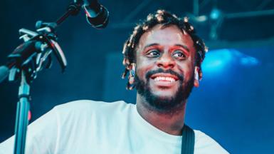 Myles Smith - a man with short dreadlocks and a short beard - smiles as he performs on stage