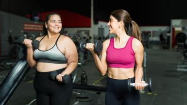 Two women in gym gear stand next to each other and lift dumbell weights. They are smiling and enjoying the workout. One of them is overweight, the other is a healthy weight and has visibly toned arms and abs. 