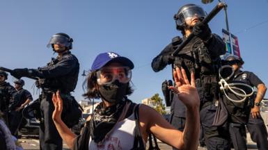 A protester wearing goggles and a face mask holds their hands in the air, while officers stand over them