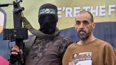 A masked Hamas fighter with Israeli hostage Eli Sharabi  on a stage before he was handed over to a Red Cross team in Deir el-Balah