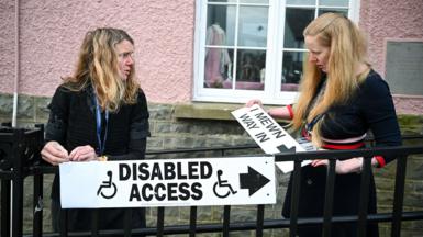 Two people erect signs outside a pink building in St Davids, Wales on Thursday morning, one saying 'disabled access' with an arrow pointing right, and the other saying 'I mewn, way in', also with an arrow.