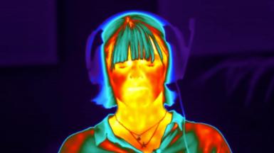 The image - taken with a thermal camera - is of a woman with closed eyes who has a set of headphones on. It is an image of a BBC science correspondent undergoing a scientific stress test whilst being filmed with a thermal camera. Her face appears red, orange and yellow - indicating warmth. While her hair and shirt and headphones appear blue, indicating that they are colder.