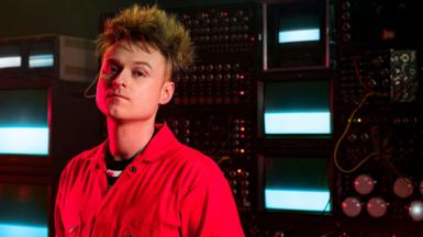 Look Mum No Computer, aka musician Sam Battle, stands in front of a bank of vintage synthesisers and TV monitors.