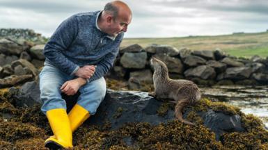 Billy Mail is sitting next to Molly the otter on rocks near the sea