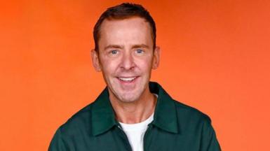 Radio presenter Scott Mills in a green shirt against an orange backdrop