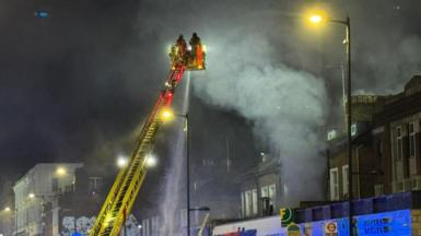 Two firefighters on a yellow turntable ladder spray water onto the fire from above. A row of shops, with homes set back from and above the shops, can be seen below