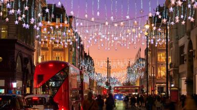 Christmas decorations light up a busy street in central London. There is a double-decker bus and pedestrians on the street.