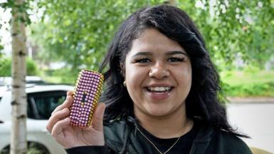 Grace, wearing a black top and jumper, smiles as she holds up her brick-phone, which she has decorated in pink and yellow plastic gems