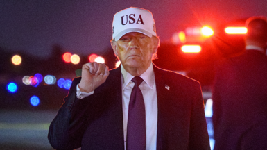 Close up of Donald Trump with his fist in the air after landing at Palm Beach international airport. It is night time and he is wearing a white cap with USA on it.